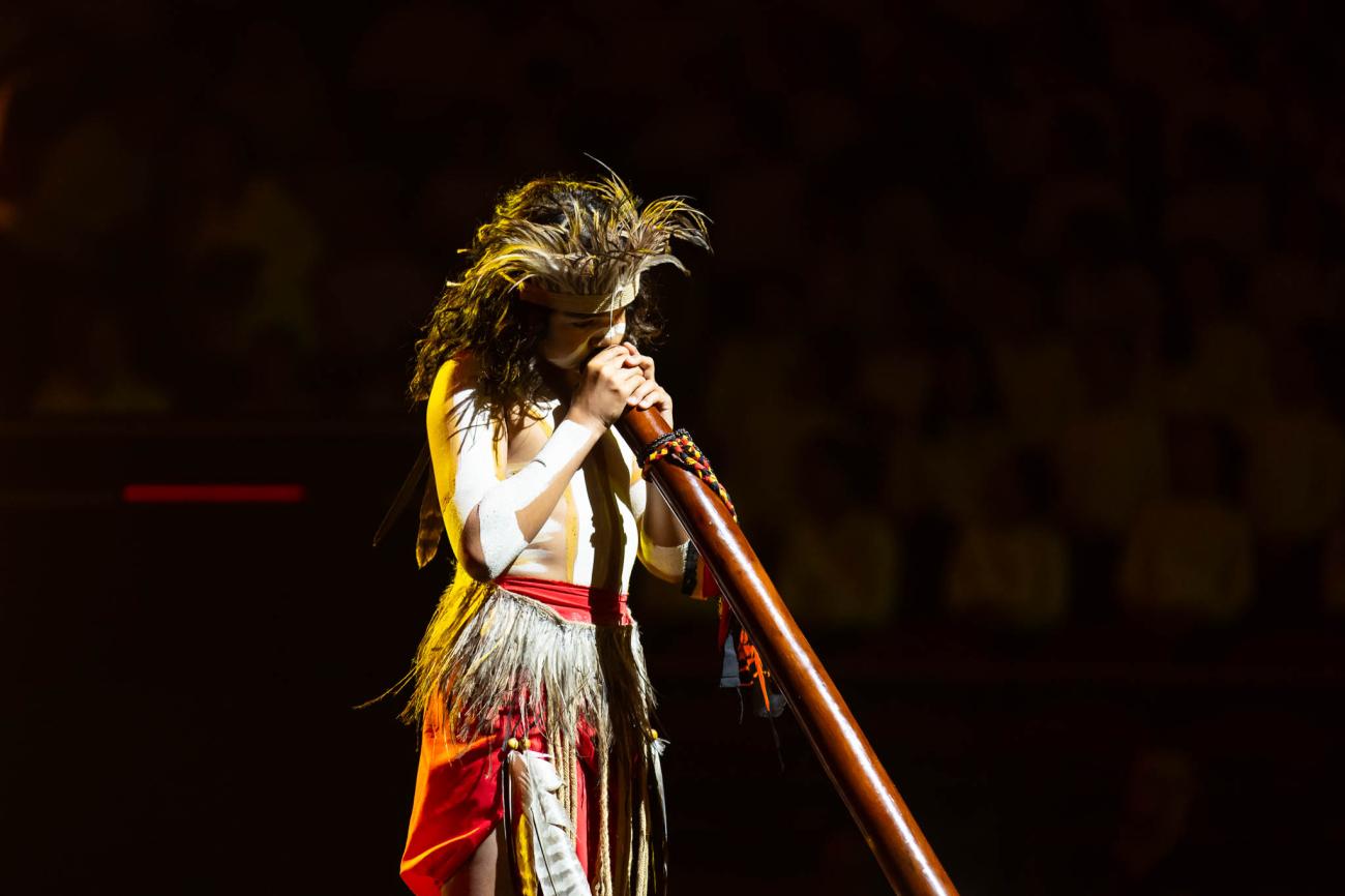 A young musician wearing feather headdress, skirt, red naga and with thick white lines of body paint, playing a didgeridoo