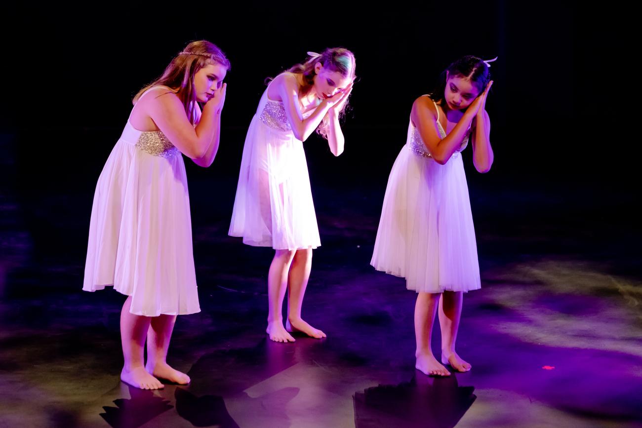 3 students on stage in white dresses with hands up at head like they are sleeping