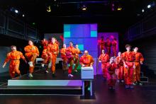 A chorus of students in bright orange jumpsuits dance on a multi-level stage