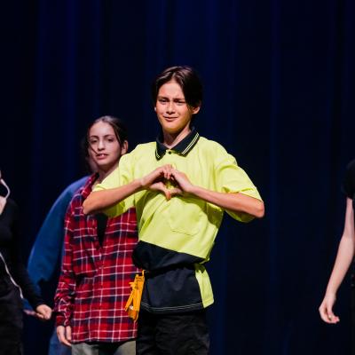 student dressed in tradie hi-vis yellow shirt makes a heart symbol with his hands while students dressed as stage managers move in the background.